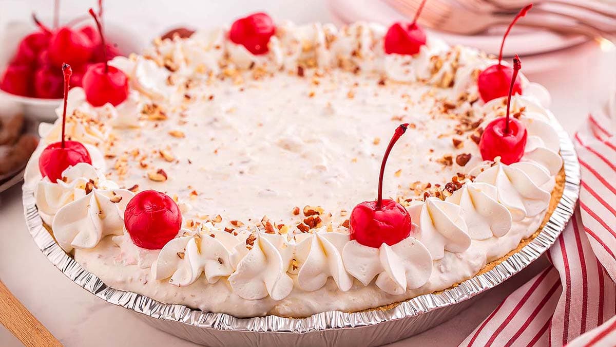 A cream pie topped with whipped cream, red cherries, and chopped nuts in a foil pie tin.