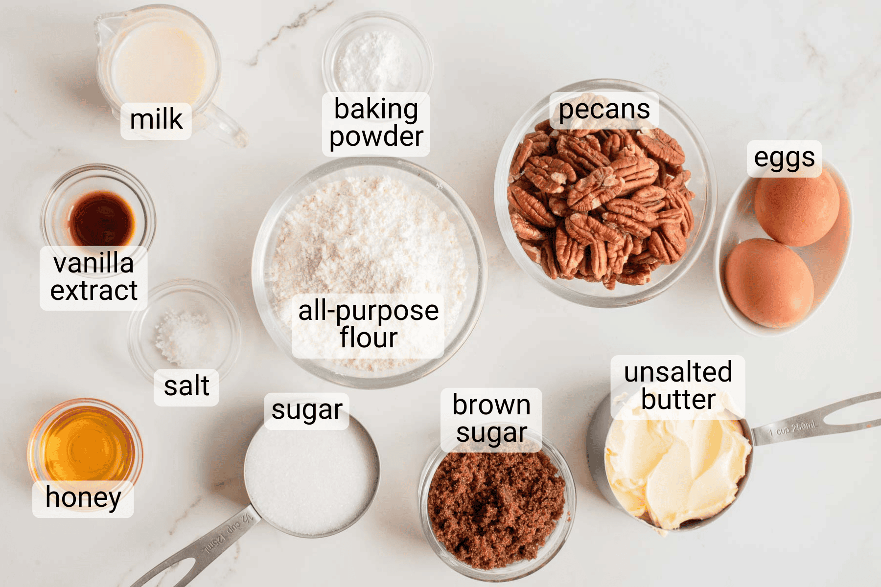 Various baking ingredients labeled in bowls and cups, including milk, baking powder, pecans, eggs, vanilla extract, salt, all-purpose flour, sugar, brown sugar, unsalted butter, and honey.