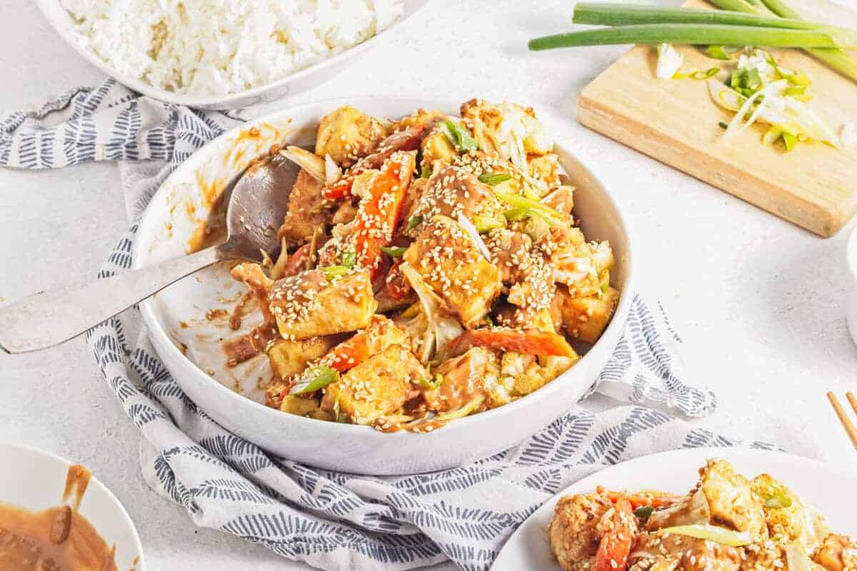 A bowl of tofu and vegetables topped with sesame seeds, served with a spoon. A side of rice and sliced green onions are visible in the background.