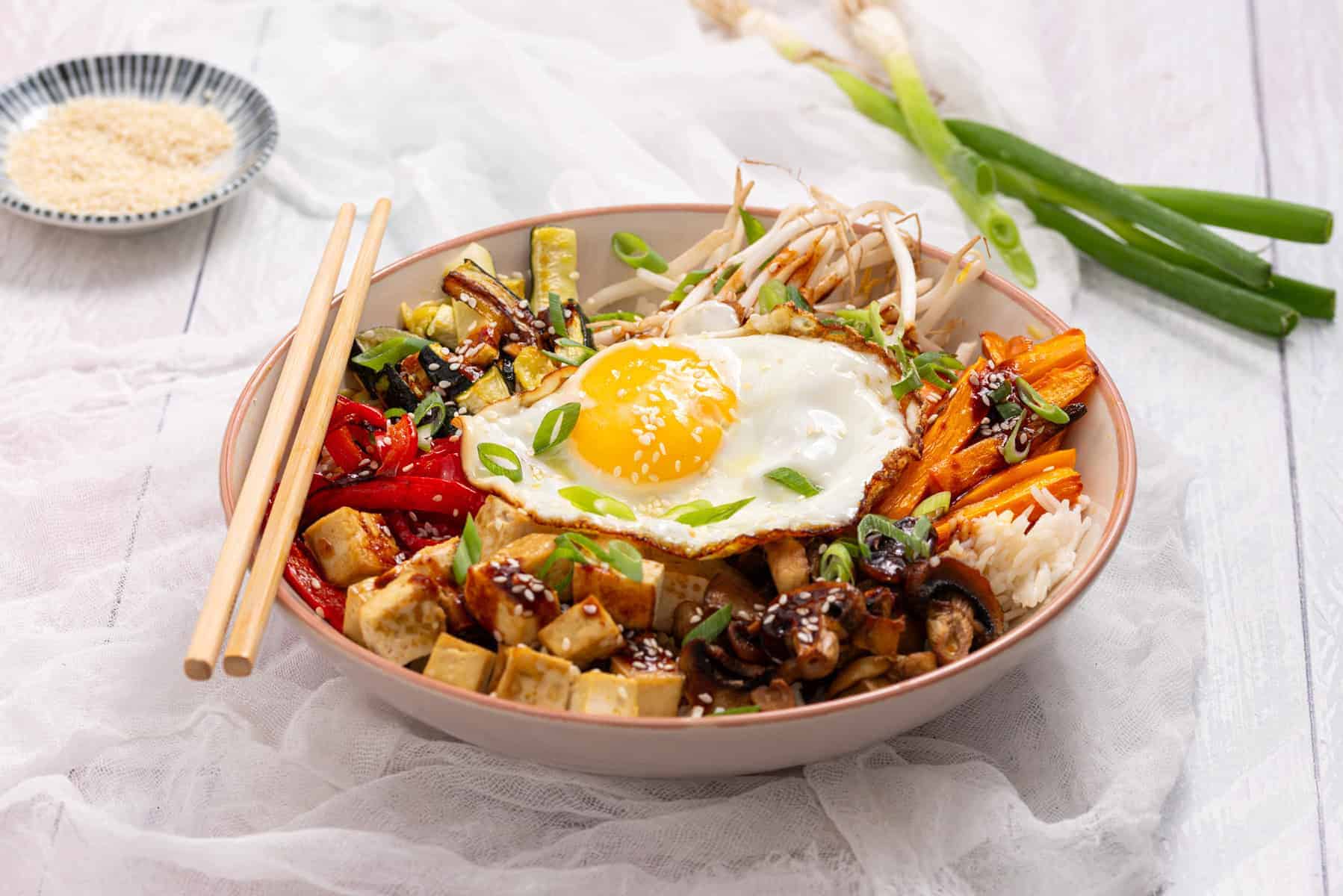 A bowl of rice topped with a sunny side up egg, tofu, sautéed vegetables, green onions, bean sprouts, and sauce, with chopsticks resting on the side.