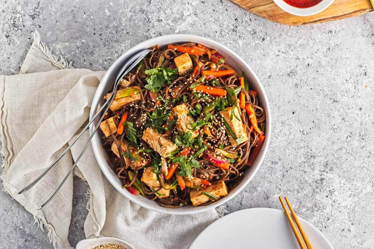 A bowl of noodle stir-fry with tofu, carrots, bell peppers, and greens, topped with sesame seeds and fresh herbs, is placed on a grey surface with a fork and napkin nearby.
