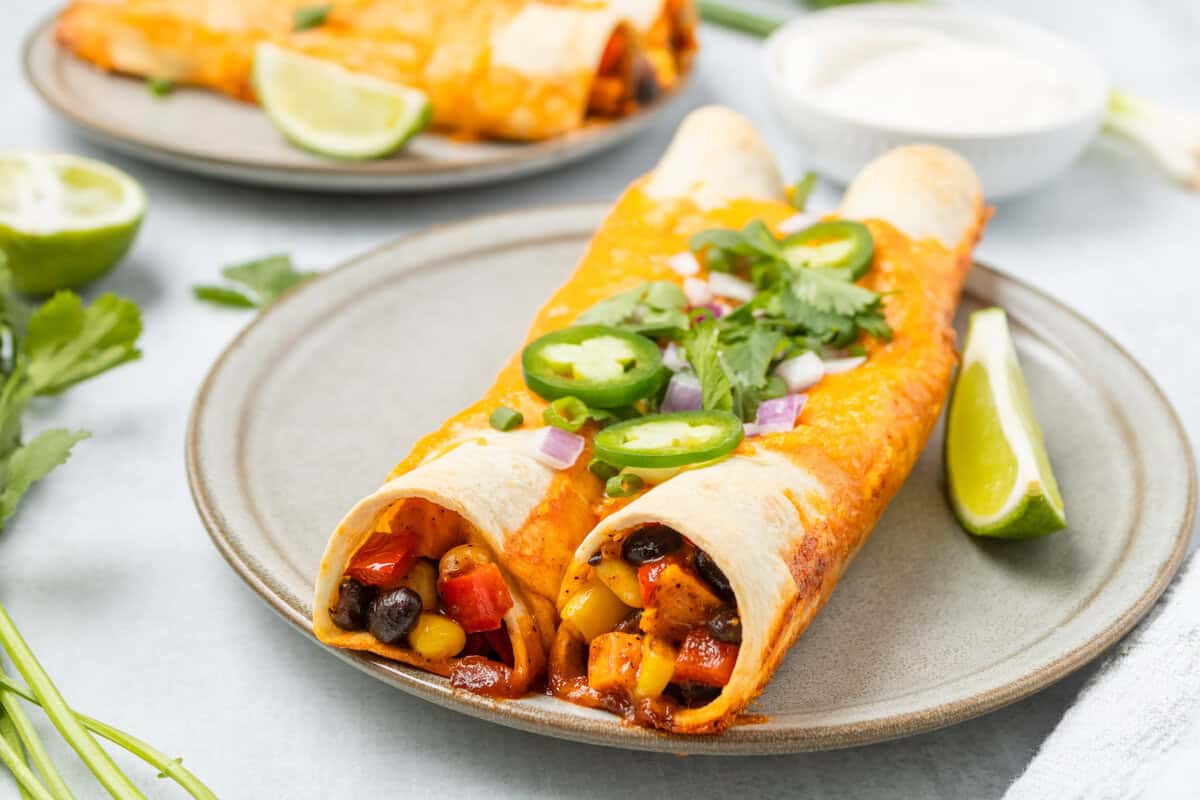 Two vegetable enchiladas topped with jalapeño slices, onions, and cilantro are served on a plate with a lime wedge; a bowl of sauce and another plate are in the background.