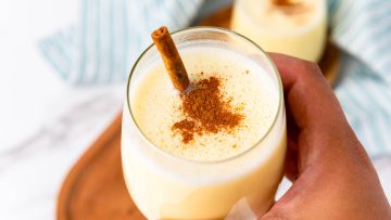 A hand holds a glass of eggnog topped with ground cinnamon and a cinnamon stick, with another glass in the background.