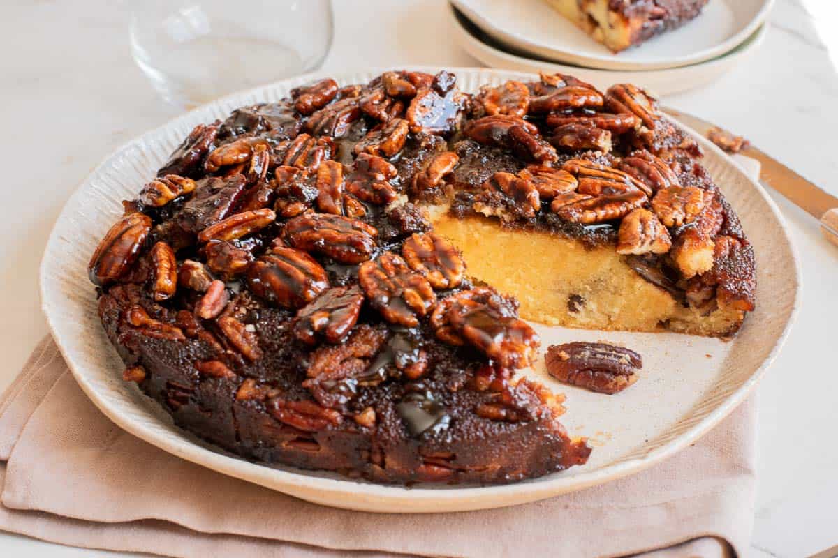 Round pecan upside-down cake with a slice missing, showing moist yellow interior and caramelized pecans on top, served on a light plate with a napkin underneath.
