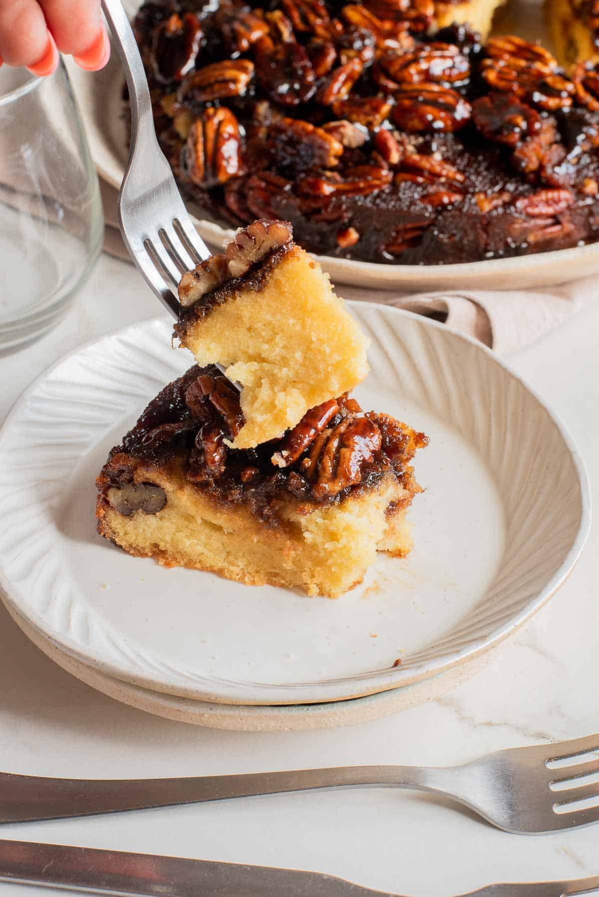 A slice of pecan upside-down cake on a white plate with a fork lifting a bite-sized piece.