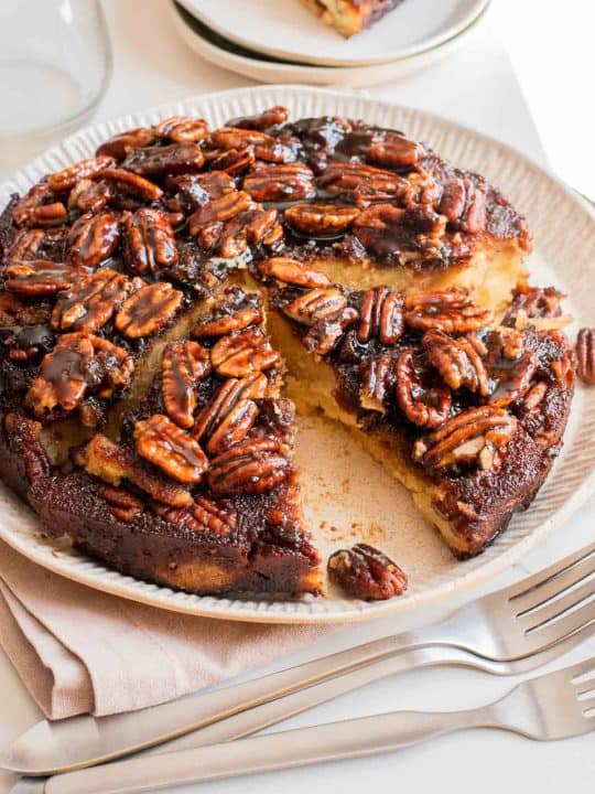 A round pecan-topped cake with a slice removed, displayed on a plate with utensils and a napkin beside it.