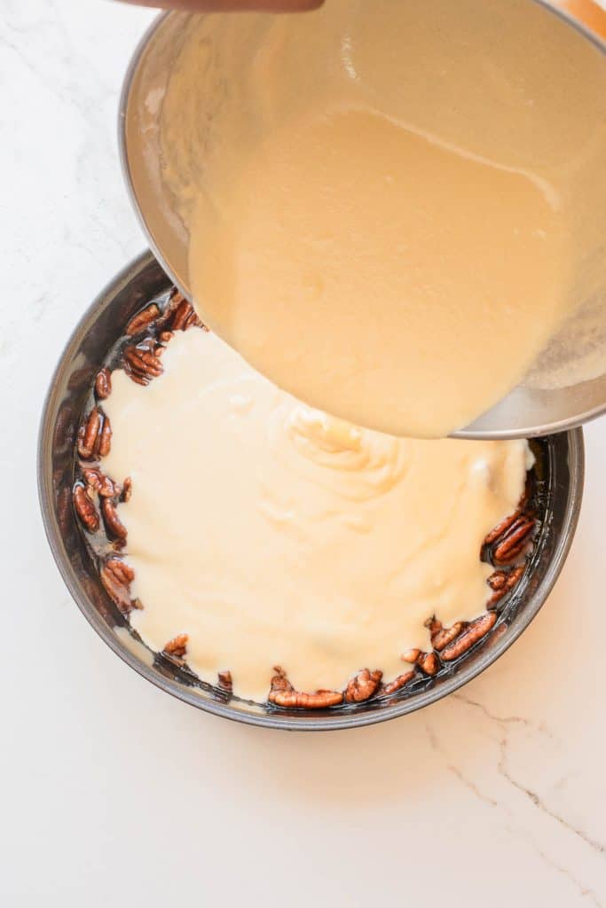 Batter is being poured from a mixing bowl into a round cake pan lined with pecans on the bottom.