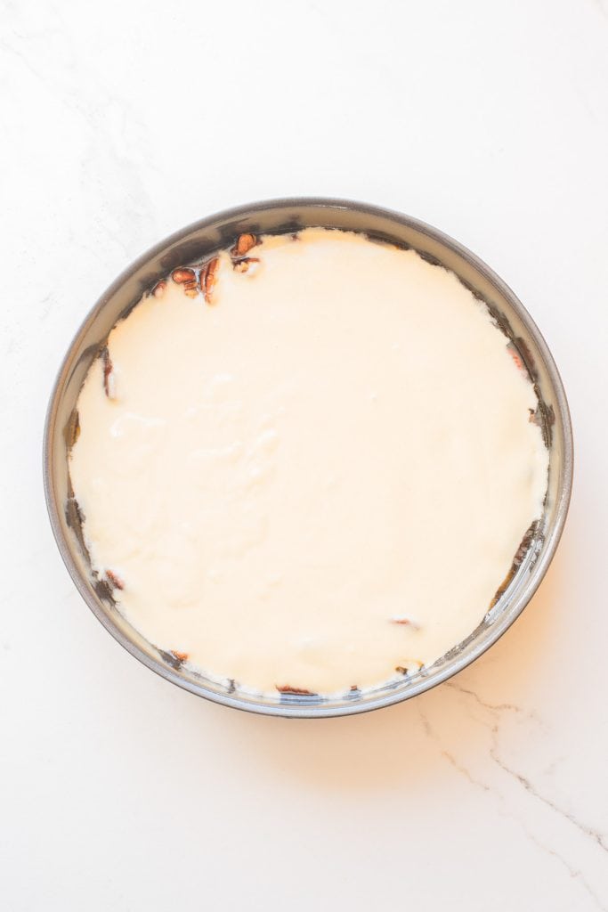 A round cake pan filled with a smooth, pale batter, partially covering pieces of pecan on the bottom, placed on a white marble surface.