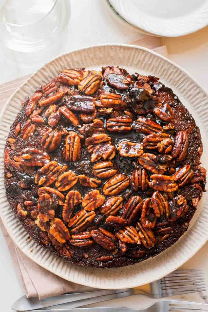 A round pecan cake with a glossy, caramelized pecan topping is served on a beige plate, with utensils and dishes nearby.