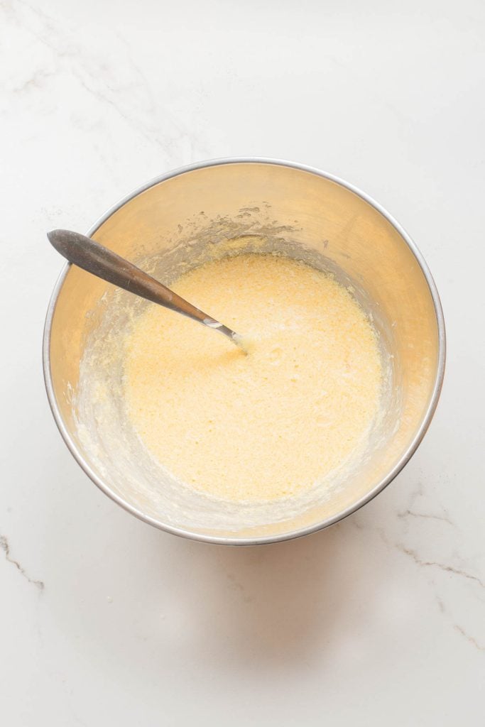 A metal mixing bowl with yellow batter and a spoon sits on a white marble surface.