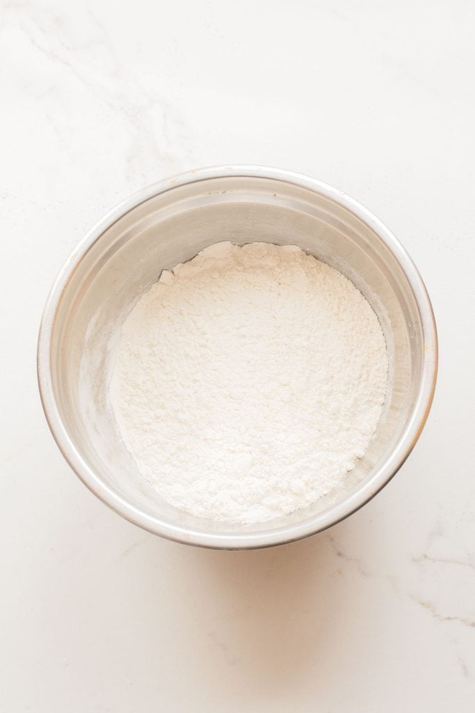 A metal mixing bowl containing a mound of white flour sits on a white marble surface.
