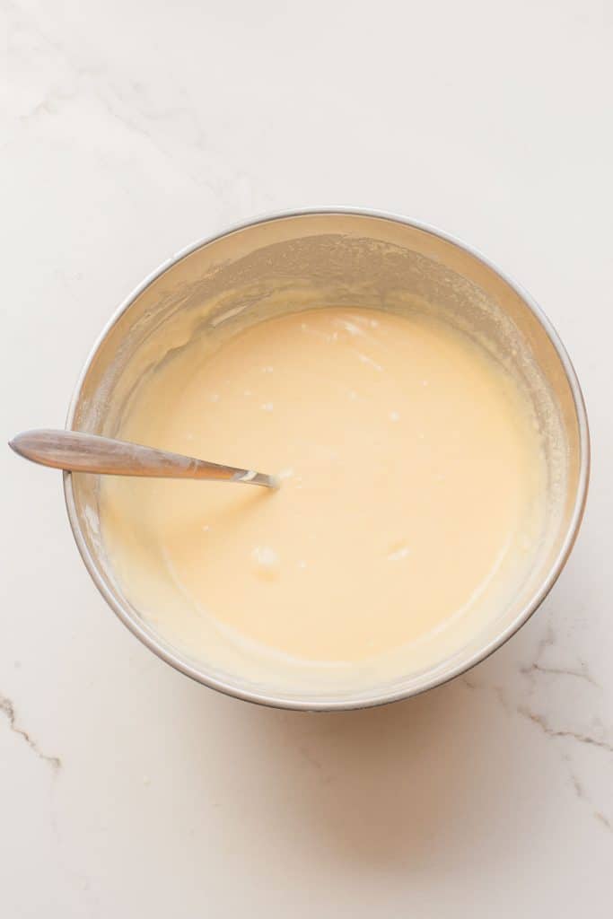 A stainless steel bowl filled with smooth, pale yellow batter and a metal spoon resting inside, placed on a light-colored surface.