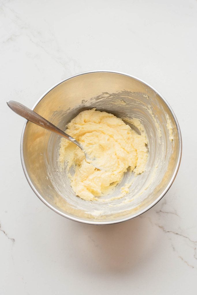 A metal mixing bowl with a spoon containing a creamy, pale yellow mixture on a white countertop.