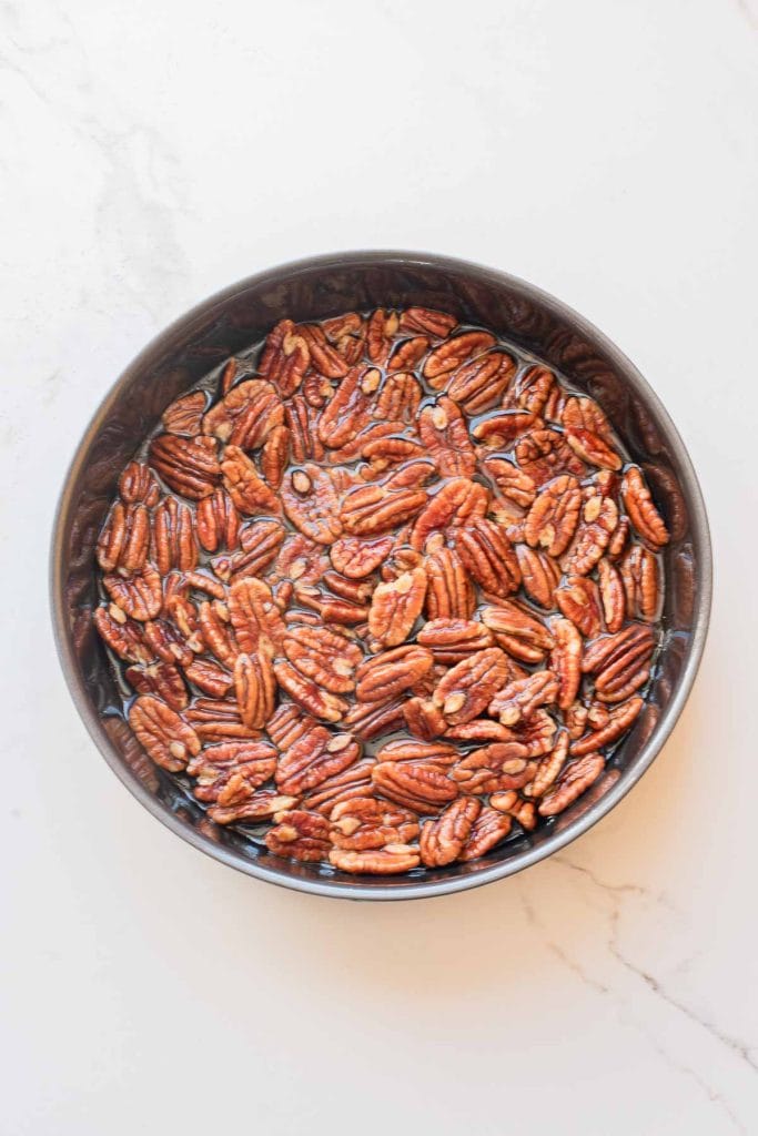 A round metal bowl filled with pecan halves soaking in water sits on a light-colored marble surface.