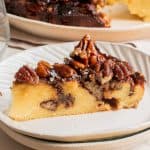A slice of pecan pie with a golden crust and pecan topping on a white plate, with more pie in the background.