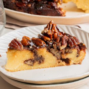 A slice of pecan pie with a golden crust and pecan topping on a white plate, with more pie in the background.