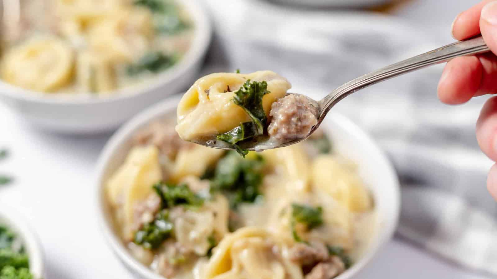A spoon holidng sausage and tortellini soup over a bowl of soup.