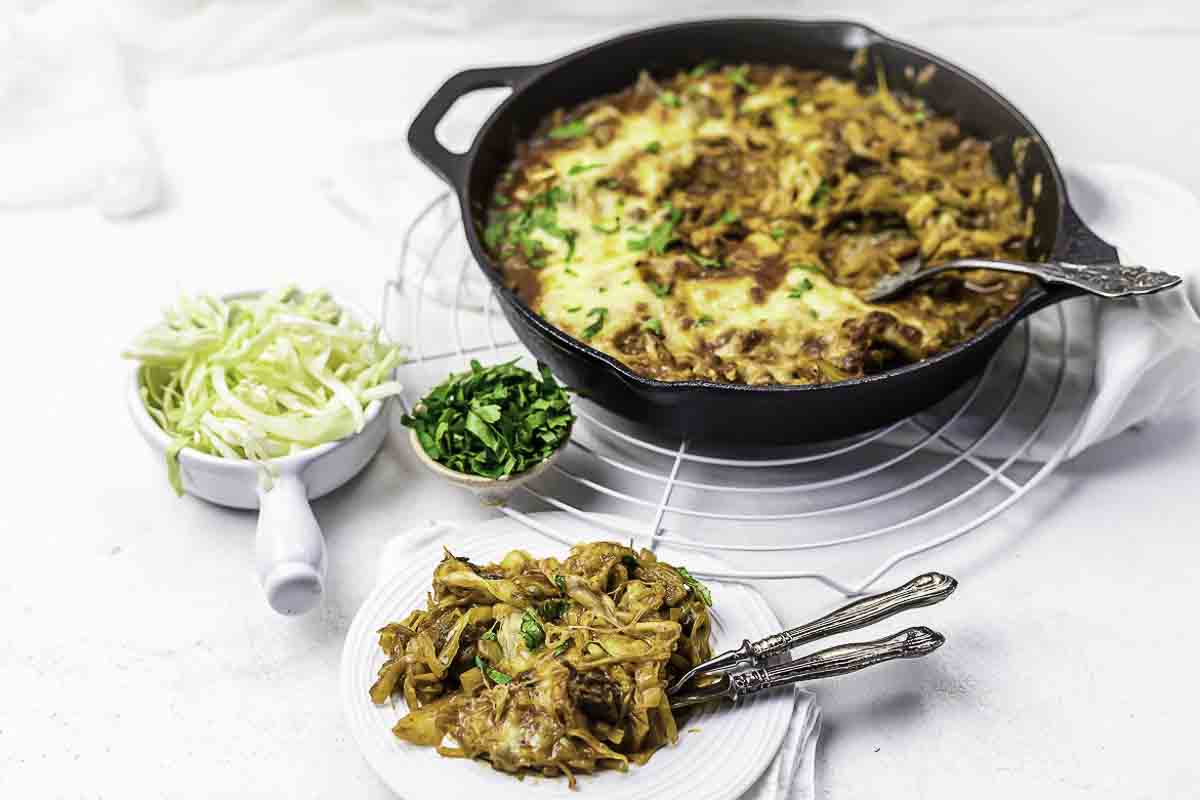A cast iron skillet filled with baked cheesy pasta, served with a portion on a plate, and small bowls of chopped parsley and shredded cabbage on the side.