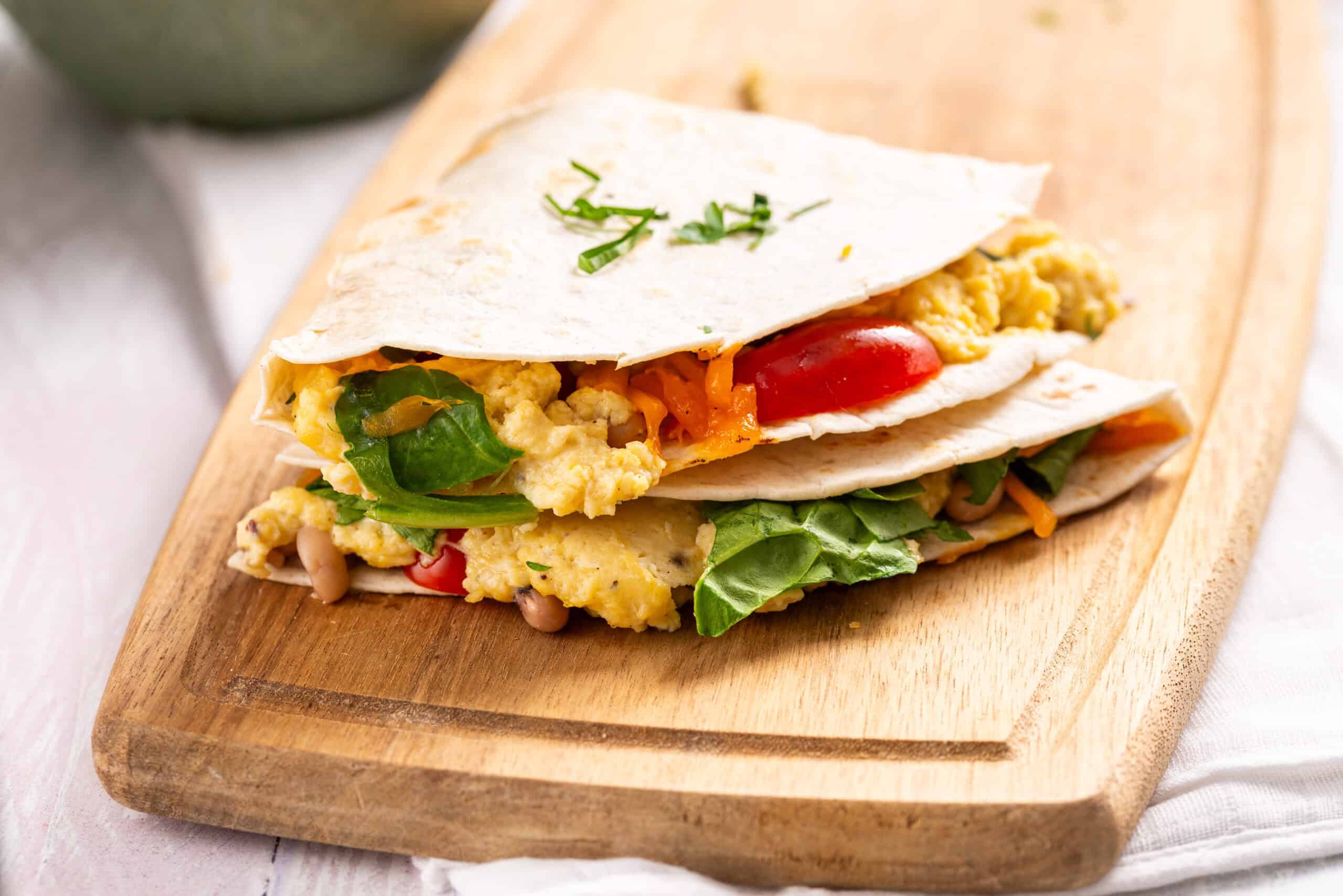 A folded tortilla filled with scrambled eggs, spinach, tomatoes, and shredded cheese on a wooden cutting board.