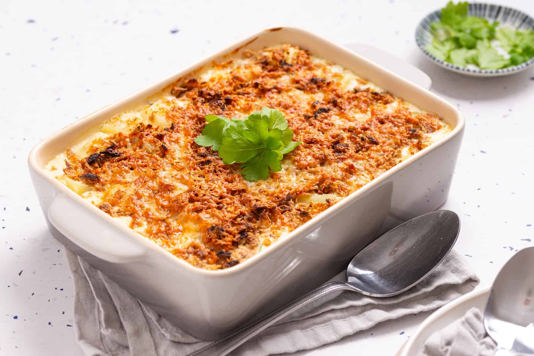 A baked casserole topped with browned cheese and garnished with fresh parsley, in a white dish beside a spoon and a small bowl of herbs.