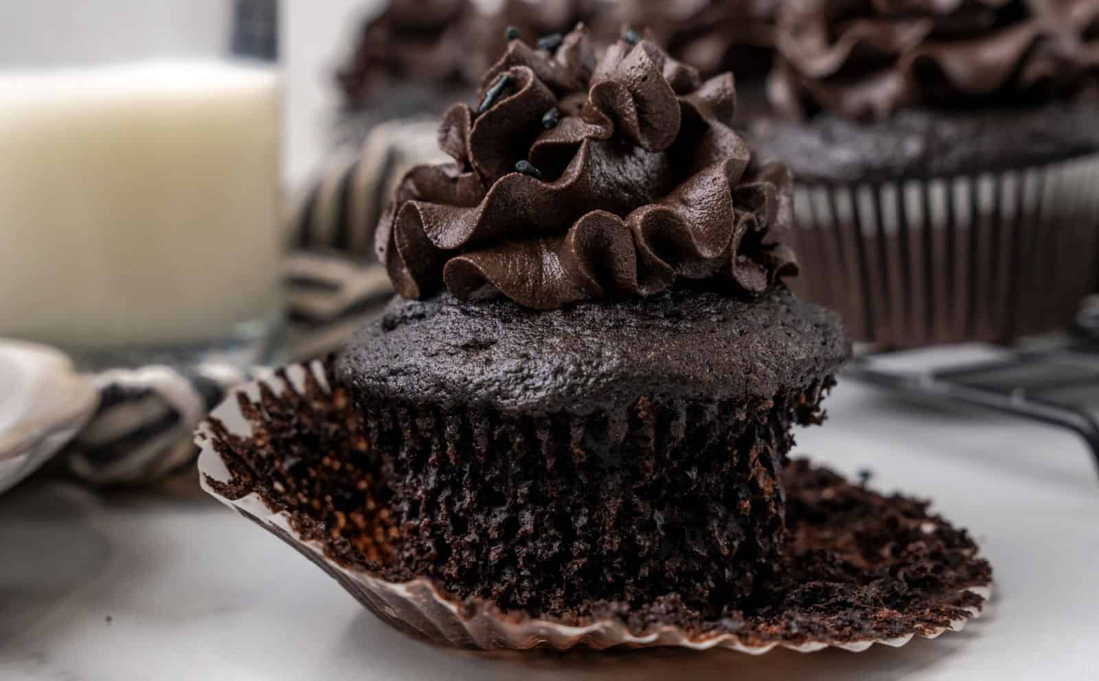 A chocolate cupcake with chocolate frosting sits on an unwrapped cupcake liner, with a bite taken out. A glass of milk and more cupcakes are in the background.