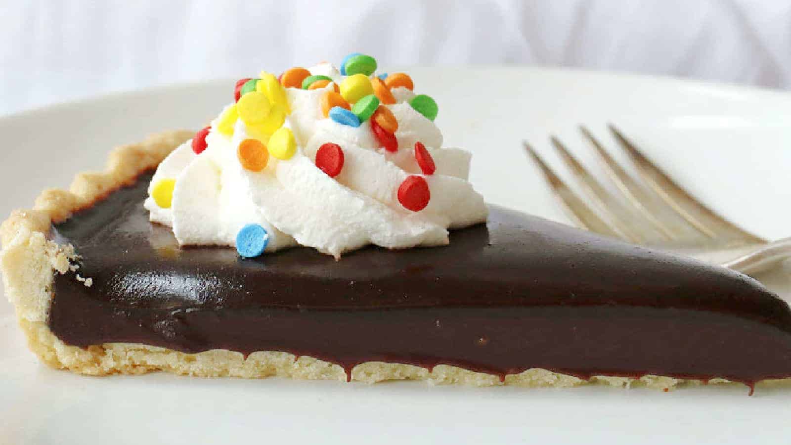 A slice of chocolate pie with a flaky crust, topped with a swirl of whipped cream and colorful sprinkles, sits on a white plate next to a fork.