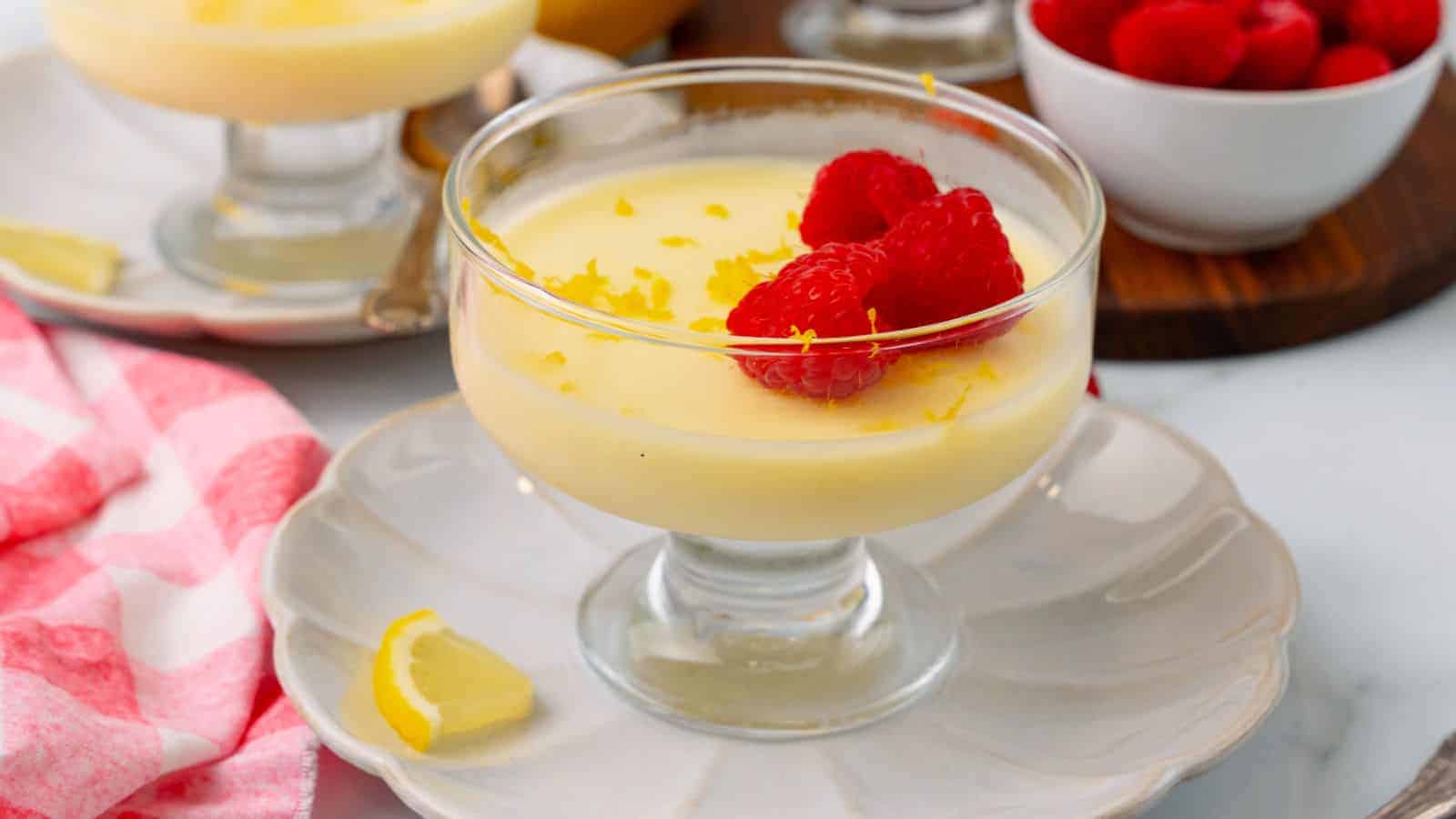 A glass dessert dish filled with creamy lemon posset, topped with fresh raspberries and lemon zest, sits on a white plate with a lemon wedge.