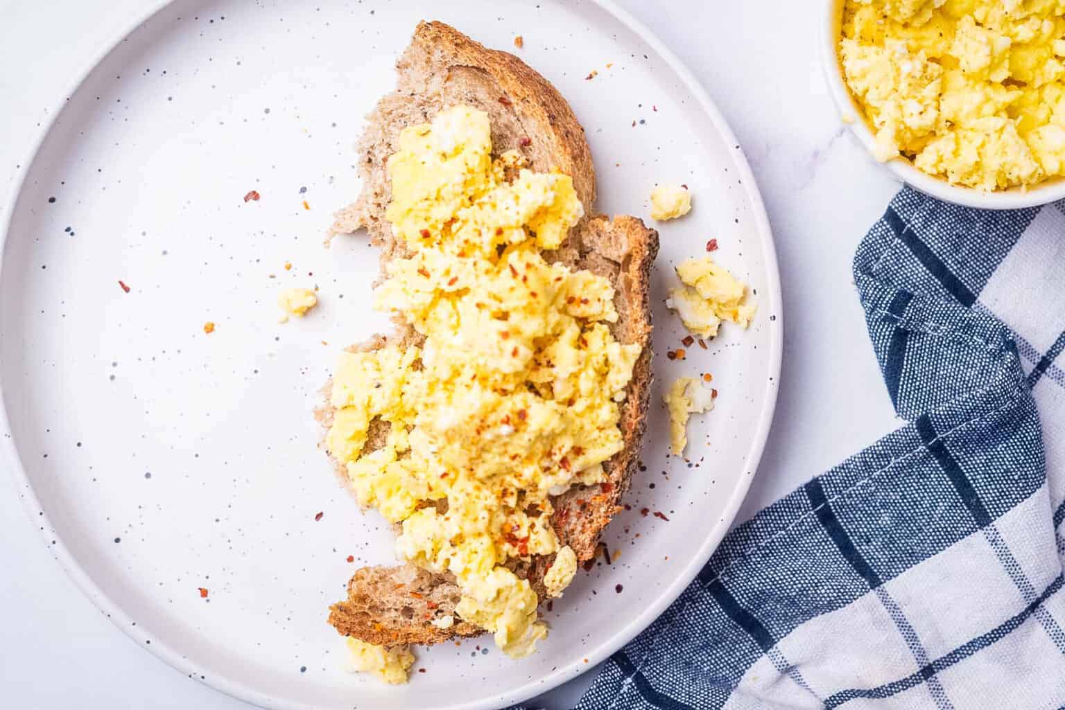 A slice of whole grain bread topped with scrambled eggs on a white plate, next to a blue checked cloth and a small bowl of scrambled eggs.