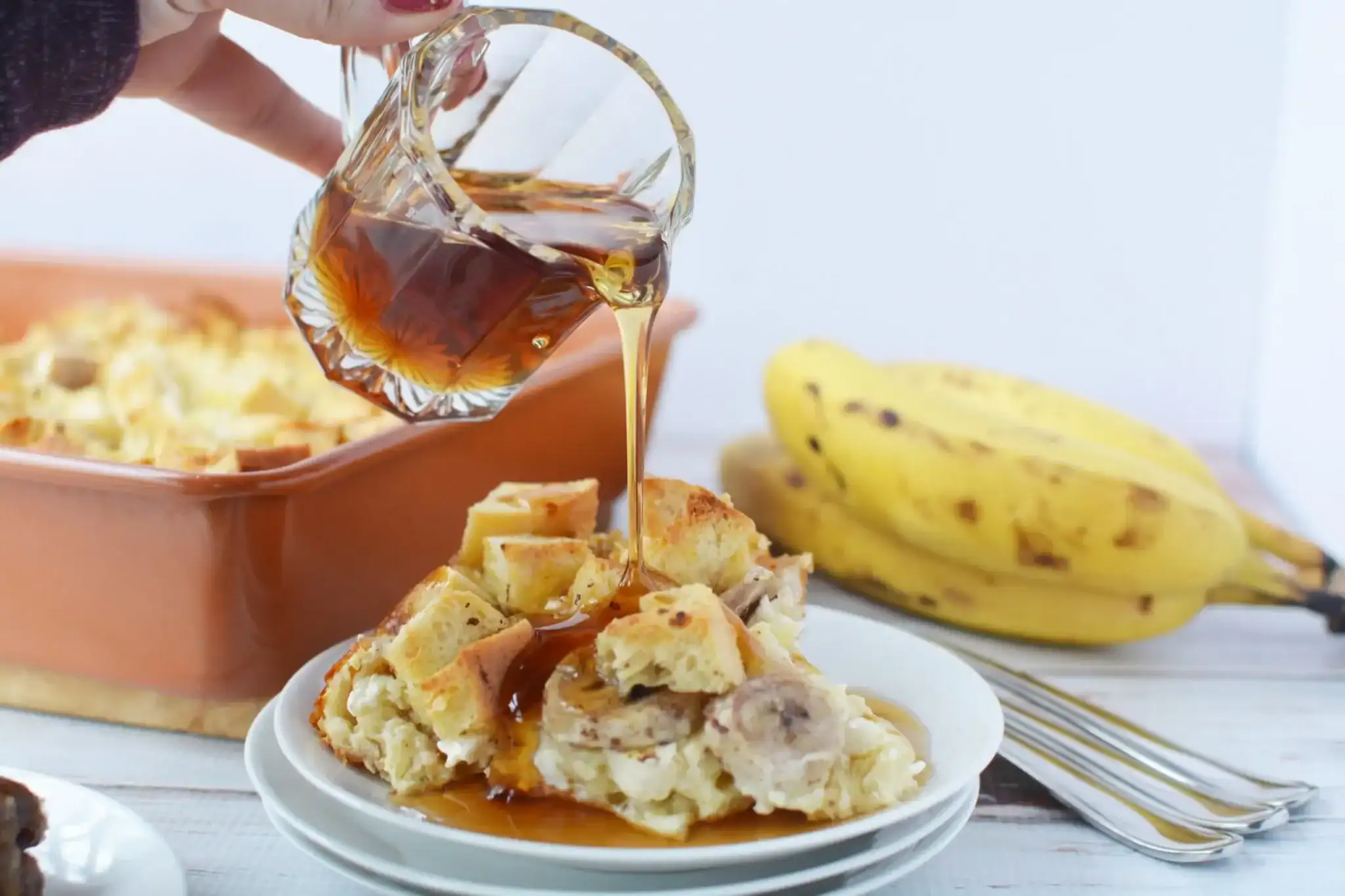 A hand pours syrup over a serving of baked casserole with bread and banana slices on a plate; a baking dish, bananas, and forks are in the background.
