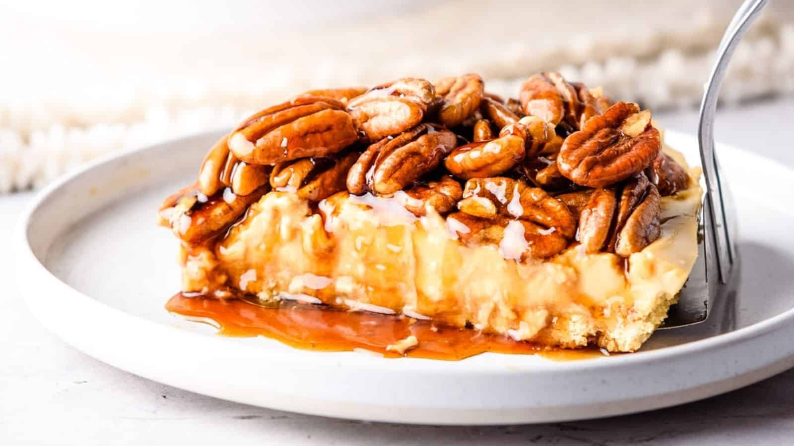 A slice of pecan-topped cheesecake with caramel sauce on a white plate, with a fork cutting into it.