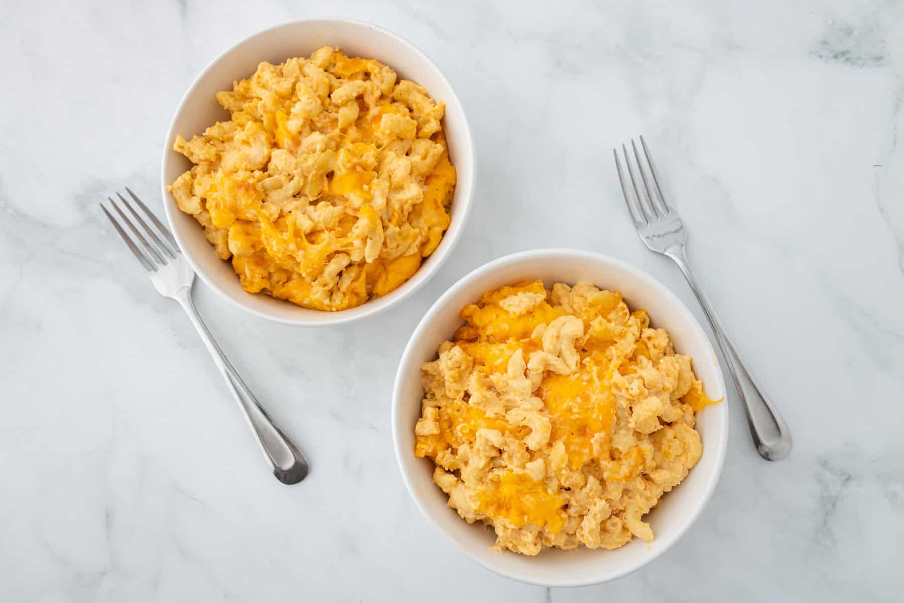 Two bowls of macaroni and cheese, each with a fork placed beside them, on a white marble surface.