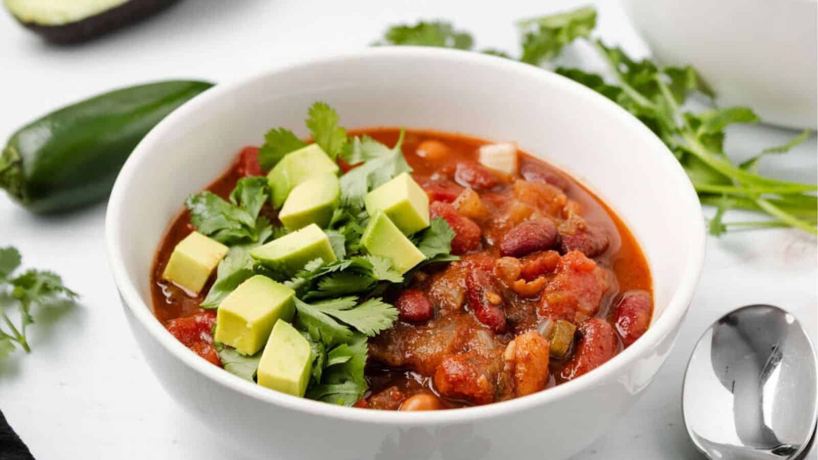 A white bowl of chili topped with diced avocado and fresh cilantro, placed on a white surface with a spoon and cilantro in the background.