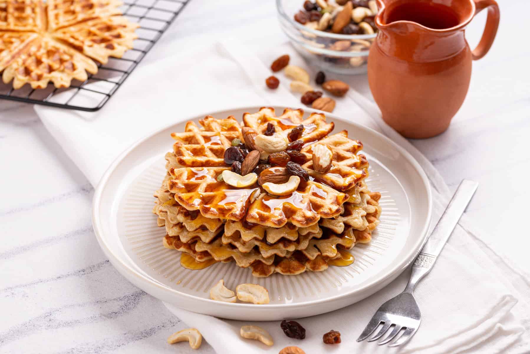 A stack of waffles topped with mixed nuts and raisins on a white plate, with a fork, cup of syrup, and extra nuts nearby.