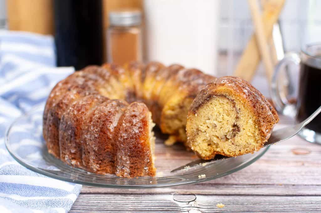 A sliced bundt cake with a cinnamon swirl sits on a glass plate; one piece is being lifted with a cake server.