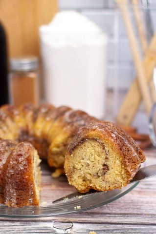 A close-up of a sliced bundt cake with a swirl pattern, resting on a glass plate with a knife, and baking ingredients visible in the background.