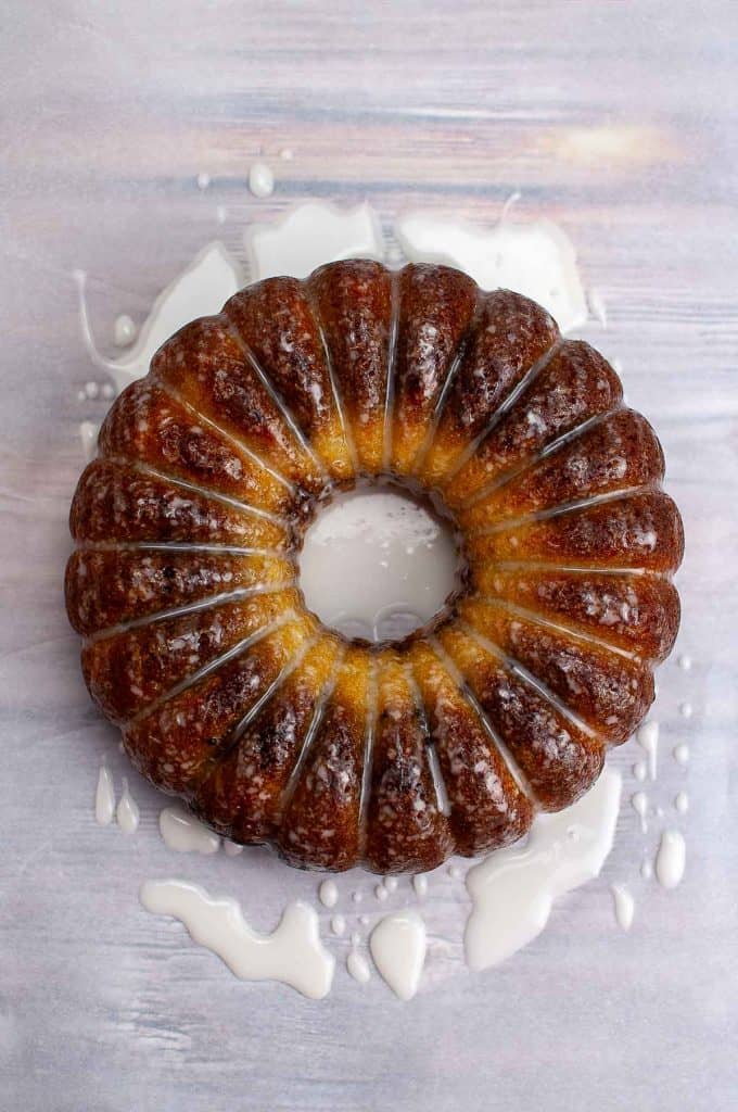 A brown Bundt cake with glaze drizzled on top and pooled around the base, displayed on a light-colored surface.