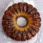 A glazed bundt cake with a golden brown exterior sits on a light surface with drizzled icing around it.