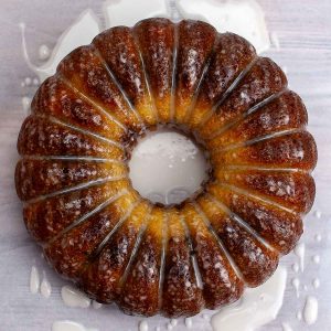 A glazed bundt cake with a golden brown exterior sits on a light surface with drizzled icing around it.
