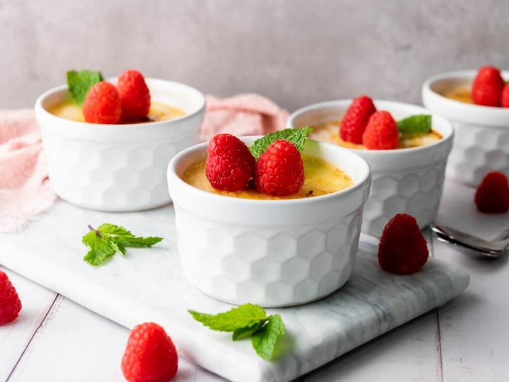 Four white ramekins of crème brûlée topped with fresh raspberries and mint leaves are arranged on a marble board, with extra raspberries and mint scattered nearby.