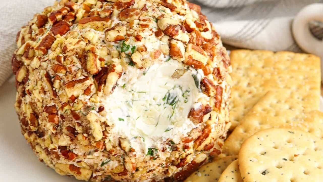 A garlic herb cheese ball coated with chopped nuts sits on a plate, surrounded by a variety of crackers. A portion of the cheeseball has been scooped out, revealing its creamy interior. A striped cloth is visible in the background