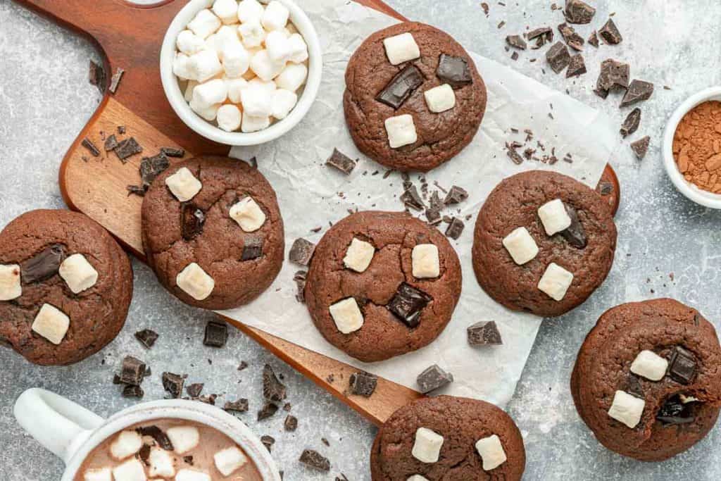 Chocolate cookies with marshmallows and chocolate chunks on a wooden board, surrounded by marshmallows, chocolate pieces, and a cup of hot cocoa.