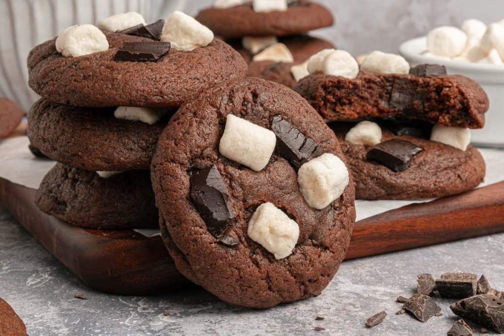 A stack of chocolate cookies topped with marshmallows and chocolate chunks sits on a wooden board, with one cookie broken in half.
