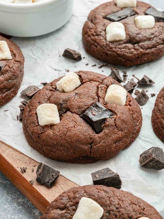 Chocolate cookies topped with chunks of dark chocolate and mini marshmallows, arranged on parchment paper with additional chocolate pieces scattered around.