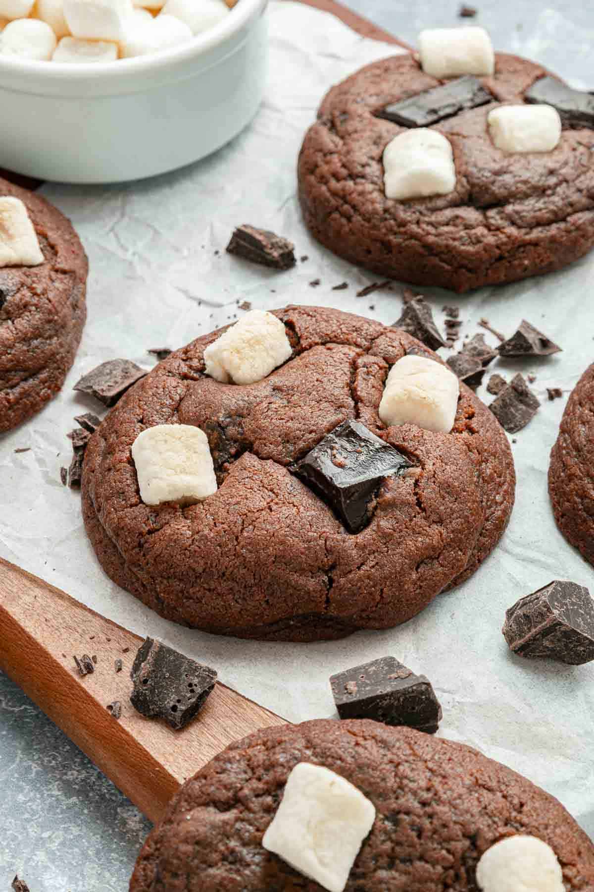 Chocolate cookies topped with chunks of dark chocolate and mini marshmallows, arranged on parchment paper with additional chocolate pieces scattered around.