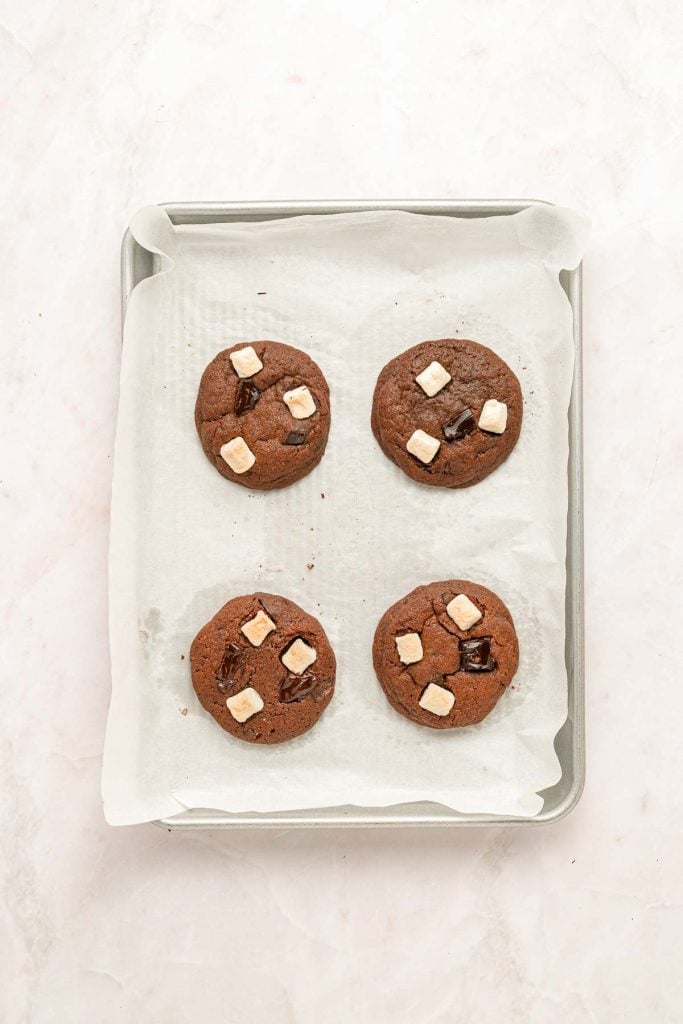 Four chocolate cookies with white and dark chocolate chunks are arranged on a parchment-lined baking sheet.