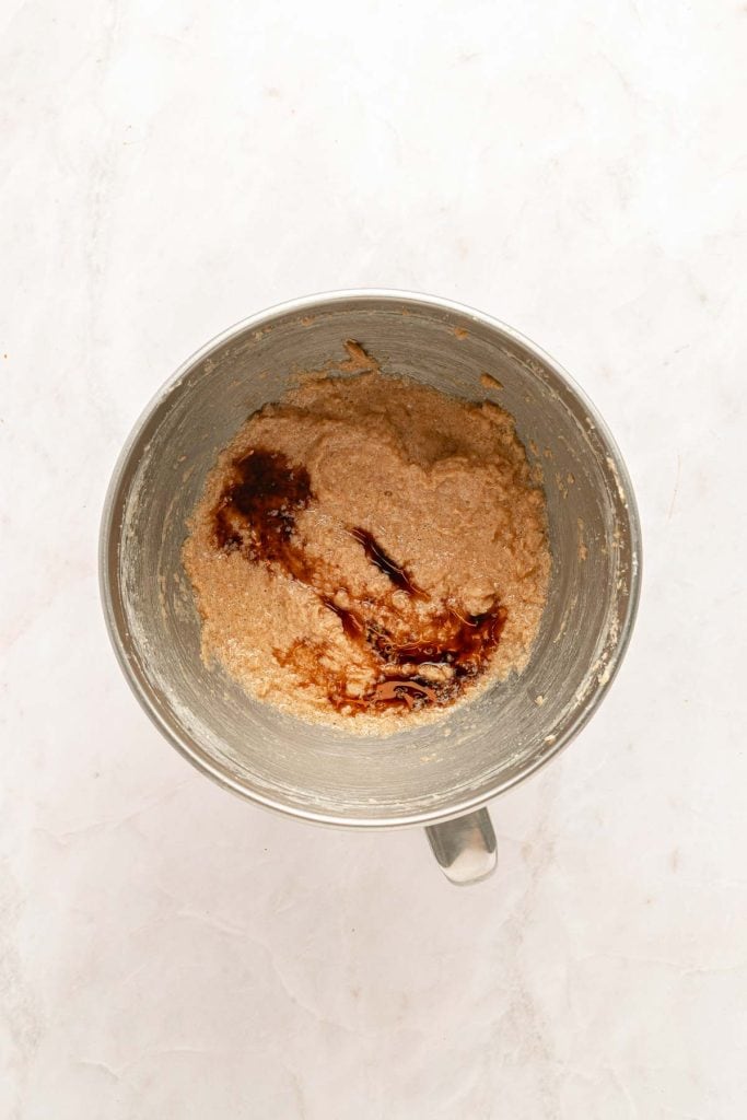 A metal mixing bowl containing partially mixed batter with dark liquid, likely vanilla extract, on a light marble surface.
