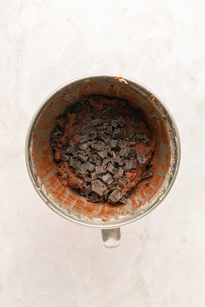 A mixing bowl containing chocolate cookie dough with chocolate chunks on top, viewed from above on a light surface.