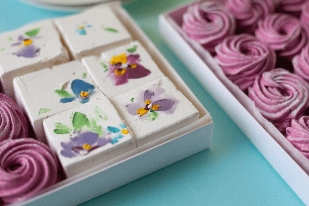 Boxes of pastel-colored marshmallow sweets, with one set decorated with floral designs and the other arranged in pink swirls, on a light blue surface.