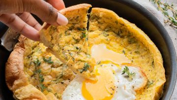 A hand lifts a slice of savory Dutch baby pancake with herbs from a skillet, topped with a fried egg and garnished with fresh thyme.