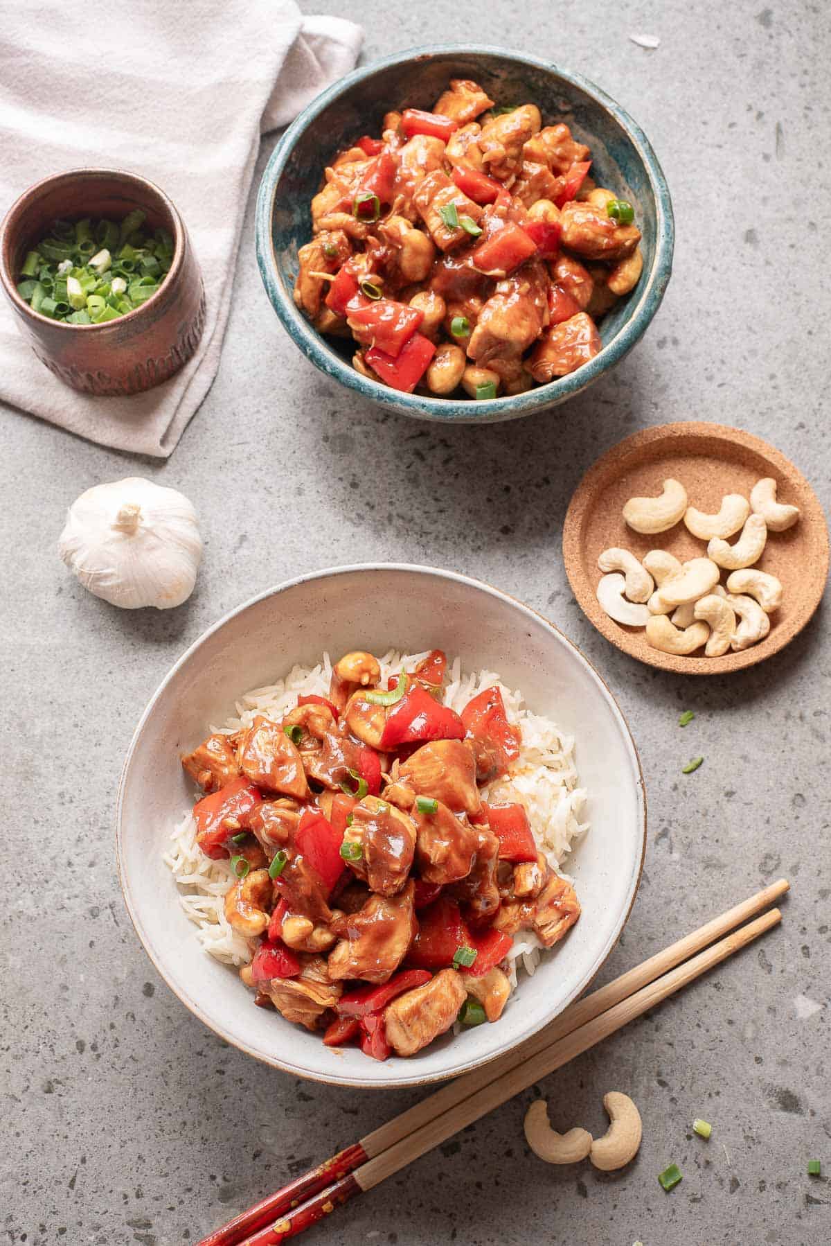 A bowl of chicken stir-fry with red bell peppers and cashews is served over rice, with extra stir-fry, cashews, green onions, and garlic arranged nearby on a gray surface.